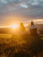 Two women sit on a grassy meadow and look towards the sunset over mountain silhouettes under a cloudy sky. | © Hochkönig Tourismus GmbH