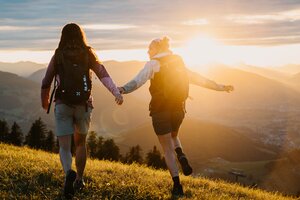 Two women with backpacks walk across a meadow in evening light overlooking mountains and valley in the Hochkönig region. | © Hochkönig Tourismus