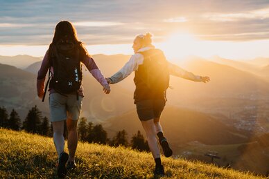 Zwei Frauen mit Rucksäcken gehen über eine Wiese im Abendlicht mit Blick auf Berge und Tal in der Region Hochkönig. | © Hochkönig Tourismus