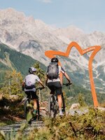 Two mountain bikers ride on a gravel trail towards an arch, with high rocky mountains in the Hochkönig region. | © Hochkönig Tourismus GmbH