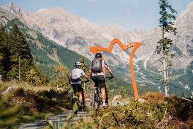 Two mountain bikers ride on a gravel trail towards an arch, with high rocky mountains in the Hochkönig region. | © Hochkönig Tourismus GmbH