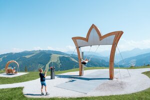 Child swings under large crown-shaped frame with Alpine view; woman takes photo; bench swing in grassy background | © Miriam Lottes