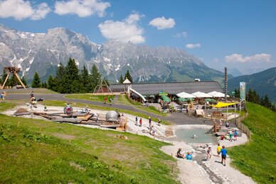 Kinder spielen an Wasserspielen und Spielgeräten auf Toni’s Almspielplatz, im Hintergrund Berge und bewirtete Hütte mit Terrasse | © Gruber Michael