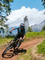 A mountain biker rides through a bend on a dirt trail between trees with mountains in the Hochkönig region. | © Hochkönig Tourismus GmbH