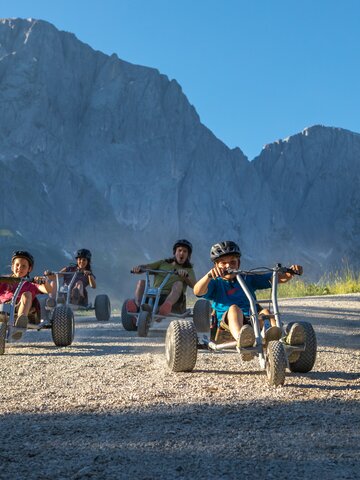 Kinder fahren mit Mountaincarts bergab vor steiler Hochkönig-Kulisse, alle tragen Helme, sonniges Wetter | © Roland Haschka