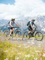 Zwei Mountainbiker fahren auf einem Schotterweg vor steilen Felsbergen und grünen Almwiesen am Hochkönig. | © Hochkönig Tourismus GmbH