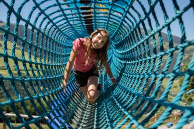 Smiling girl crawls through blue rope tunnel at Castle Playground Natrun, alpine meadow in background | © Roland Haschka