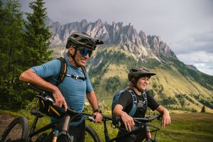 Two mountain bikers stand with bikes in front of steep rocky peaks and green slopes in the Hochkönig region. | © Hochkönig Tourismus