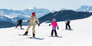 Erwachsene fährt mit drei Kindern auf breiter Piste bei Sonnenschein in den Alpen | © Hochkönig Tourismus GmbH