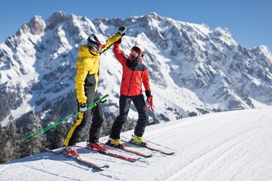 Two skiers in colourful outfits give high five on ski slope with mountain backdrop | © Hochkönig Tourismus GmbH