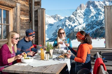Vier Personen essen und trinken auf Holzterrasse einer Berghütte mit Blick auf Alpen | © Hochkönig Tourismus GmbH