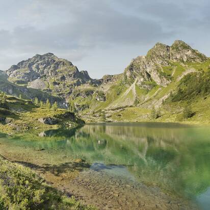Zwei Wanderer gehen entlang eines türkisgrünen Bergsees, umgeben von Almwiesen, Bäumen und felsigen Bergen | © Peter Burgstaller