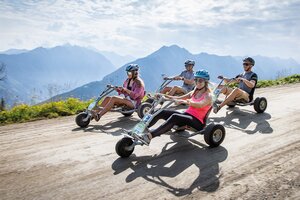 Vier Personen mit Helmen fahren auf Mountain Go-Karts eine breite Schotterstraße entlang, Berge im Hintergrund | © Planai, Harald Steiner