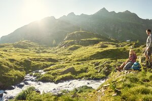 Zwei Wanderer rasten an einem Gebirgsbach in der Sonne mit grünem Almgelände und Bergen im Hintergrund | © Peter Burgstaller
