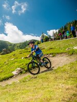 Ein Kind mit Helm fährt auf einem Mountainbike einen kurvigen Trail über eine grüne Alm, Menschen im Hintergrund | © Christine Höflehner