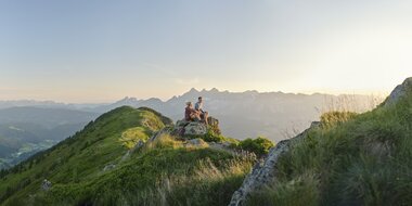 Zwei Wanderer sitzen auf einem Felsen und blicken beim Sonnenaufgang über die Berge der Reiteralm | © Peter Burgstaller