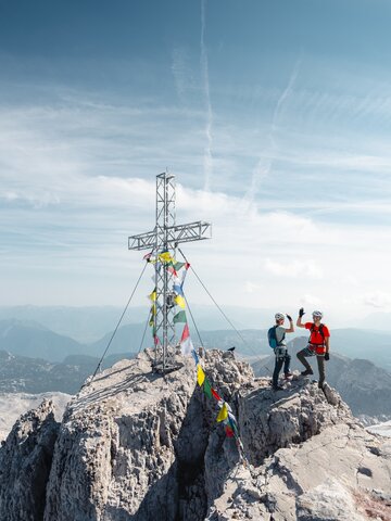 Zwei Kletterer mit Helmen stehen neben einem Gipfelkreuz auf felsigem Berggipfel über weiter Alpenlandschaft | © Mathäus Gartner