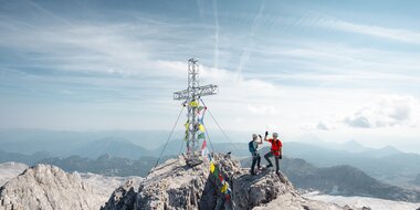 Zwei Kletterer mit Helmen stehen neben einem Gipfelkreuz auf felsigem Berggipfel über weiter Alpenlandschaft | © Mathäus Gartner