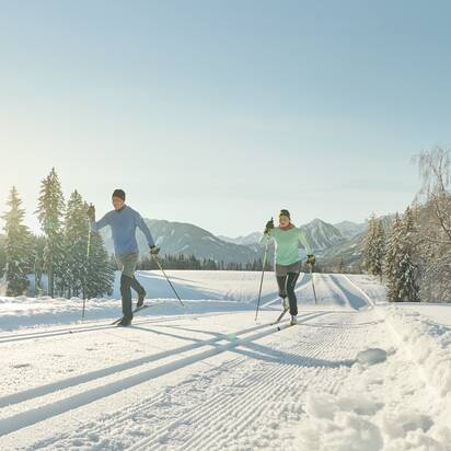 Two cross-country skiers in sportswear glide on sunny groomed trail through snowy winter landscape | © Peter Burgstaller