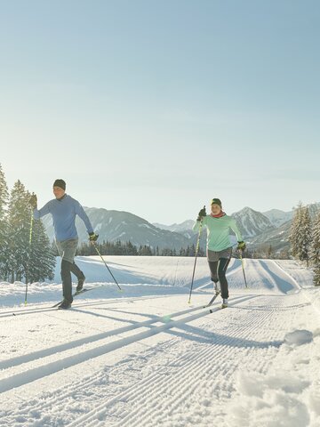 Zwei Langläufer in Sportkleidung gleiten bei Sonne auf gespurter Loipe durch verschneite Winterlandschaft | © Peter Burgstaller