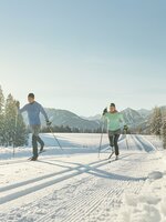 Zwei Langläufer in Sportkleidung gleiten bei Sonne auf gespurter Loipe durch verschneite Winterlandschaft | © Peter Burgstaller