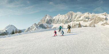Zwei Erwachsene und zwei Kinder fahren Ski vor verschneitem Dachsteinmassiv in den Alpen | © Peter Burgstaller