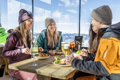 Vier Personen essen in Skikleidung an einem Holztisch mit Bergblick im Restaurant | © Mirja Geh