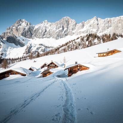 Fußspuren führen zu tief verschneiten Almhütten unterhalb des Dachsteins in winterlicher Berglandschaft | © Mathäus Gartner