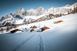 Fußspuren führen zu tief verschneiten Almhütten unterhalb des Dachsteins in winterlicher Berglandschaft | © Mathäus Gartner