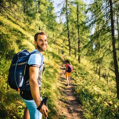 Ein Mann mit Rucksack wandert bei Sonnenschein durch einen lichten Bergwald, schaut lächelnd zurück, im Hintergrund eine Frau | © Salzburger Sportwelt - Christian Schartner