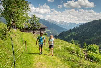 Ein Paar wandert auf einem grünen Almweg in der Salzburger Sportwelt, umgeben von Hütten und Bergen, unter blauem Himmel. | © Lorenz Masser