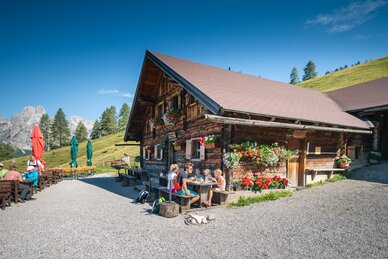 Eine Familie sitzt an einem Holztisch vor einer blumengeschmückten Alm in Filzmoos, umgeben von Bergen und grünen Wiesen. | © Salzburger Sportwelt/Christian Schartner