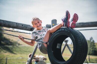 Smiling girl sits on a tire swing at the playground, holding onto chains, with grassy summer landscape in the background | © Salzburger Sportwelt