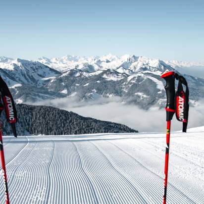 Zwei Skistöcke und rote Ski stehen auf frisch präparierter Skipiste mit Blick auf verschneite Alpenlandschaft | © Snow Space Salzburg Christian Schartner