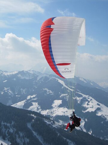 Paragleiter mit rot-blau-weißem  Schirm fliegt über von Wald und schneereichen Pisten bedeckte Berge