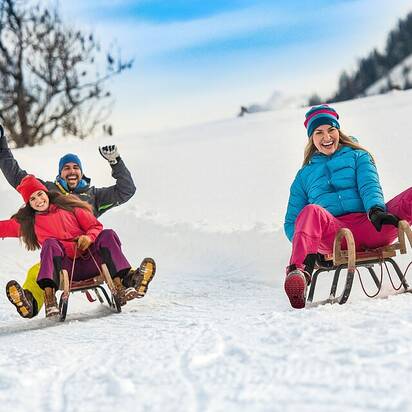 Three people joyfully sledding on a groomed snow track, surrounded by a winter landscape. | © Flachau Tourismus