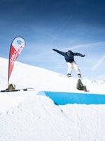 Ein Snowboarder springt bei Cash4Tricks über ein blaues Tube-Feature vor blauem Himmel, Eventflagge und Ausrüstung daneben.