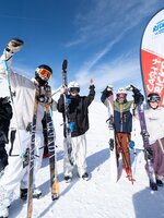 Vier junge Freeskier jubeln vor einer Cash4Tricks-Flagge im Schnee, Ski in der Hand, blauer Himmel und Berge im Hintergrund.