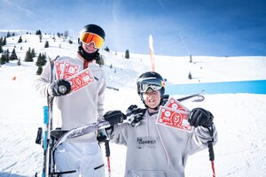 Zwei junge Freeskier posieren mit roten Cash4Tricks-Gutscheinen vor Schneekulisse, Ski und Parkelemente im Hintergrund.