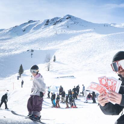 Ein Judge kniet im Schnee mit roten Cash4Tricks-Gutscheinen, viele Zuschauer bei strahlendem Sonnenschein.