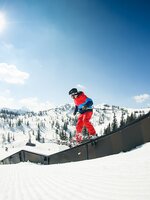 Freeskier in rot-blauem Outfit slidet auf Rail im Snowpark, Sonnenlicht und verschneite Berge bilden die Kulisse.