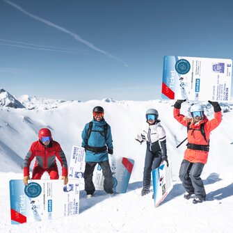 Vier Wintersportler posieren mit übergroßen Skipässen im Schnee vor alpiner Bergkulisse.