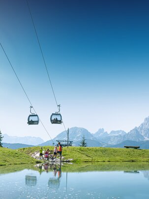 Familie am Ufer eines kleinen Bergsees unter einer Kabinenbahn, mit grünen Wiesen und Alpen im Hintergrund