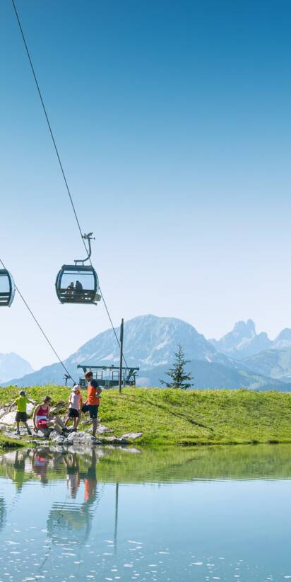 Familie am Ufer eines kleinen Bergsees unter einer Kabinenbahn, mit grünen Wiesen und Alpen im Hintergrund