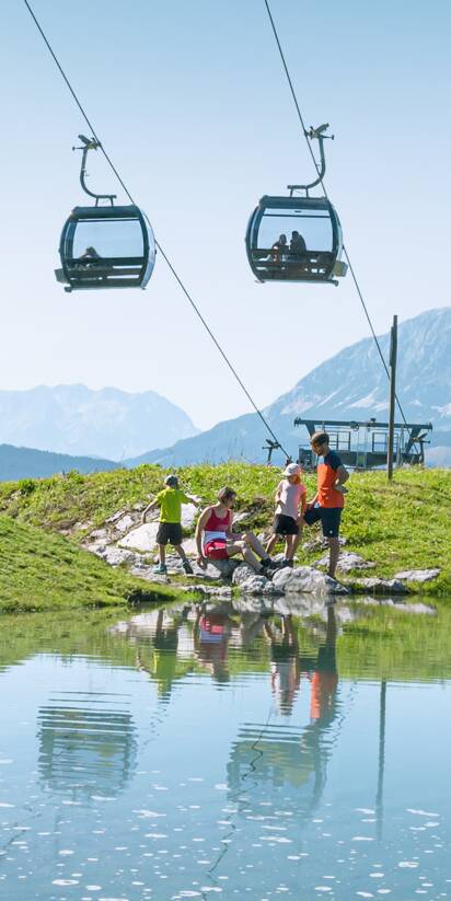 Familie am Ufer eines kleinen Bergsees unter einer Kabinenbahn, mit grünen Wiesen und Alpen im Hintergrund