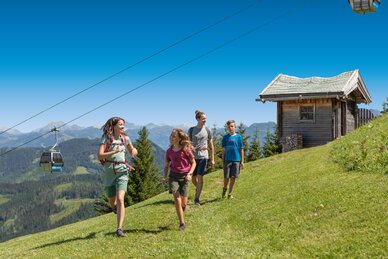 Group of four hikers enjoys a sunny day on the alpine meadow with gondola view and wooden hut in the background in Ski amadé.