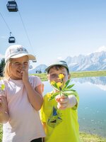 Two kids holding picked flowers at a mountain lake in Ski amadé, with the striking Dachstein mountains in the background.