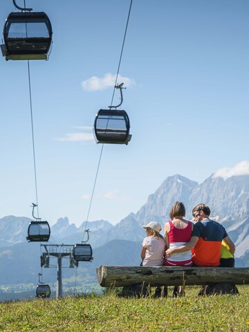 Eine Familie sitzt auf einer Holzbank unter schwebenden Gondeln und blickt auf das Panorama der steilen Berge von Ski amadé.