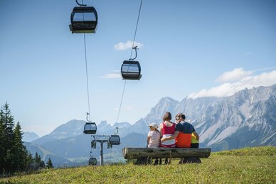 Eine Familie sitzt auf einer Holzbank unter schwebenden Gondeln und blickt auf das Panorama der steilen Berge von Ski amadé.