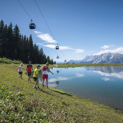 A family walks along a crystal-clear mountain lake under gondolas, enjoying the stunning view of the Dachstein mountains.